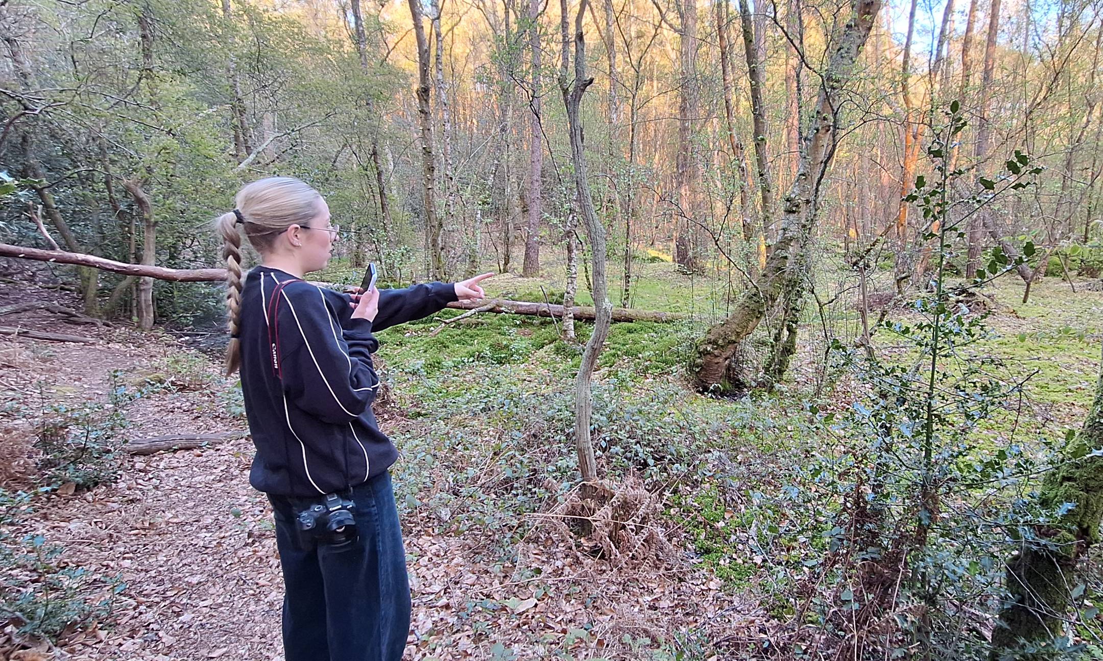 A young birdwatcher in woodland, phone in hand and camera on a strap, pointing towards a bird in the trees